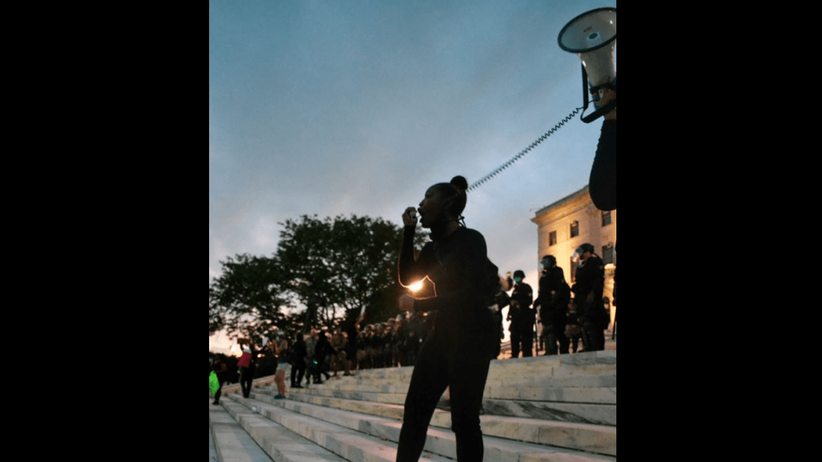 Shaffany Terrell yells into a megaphone on the steps of the Rhode Island State House. A line of State Police officers stands in the background.