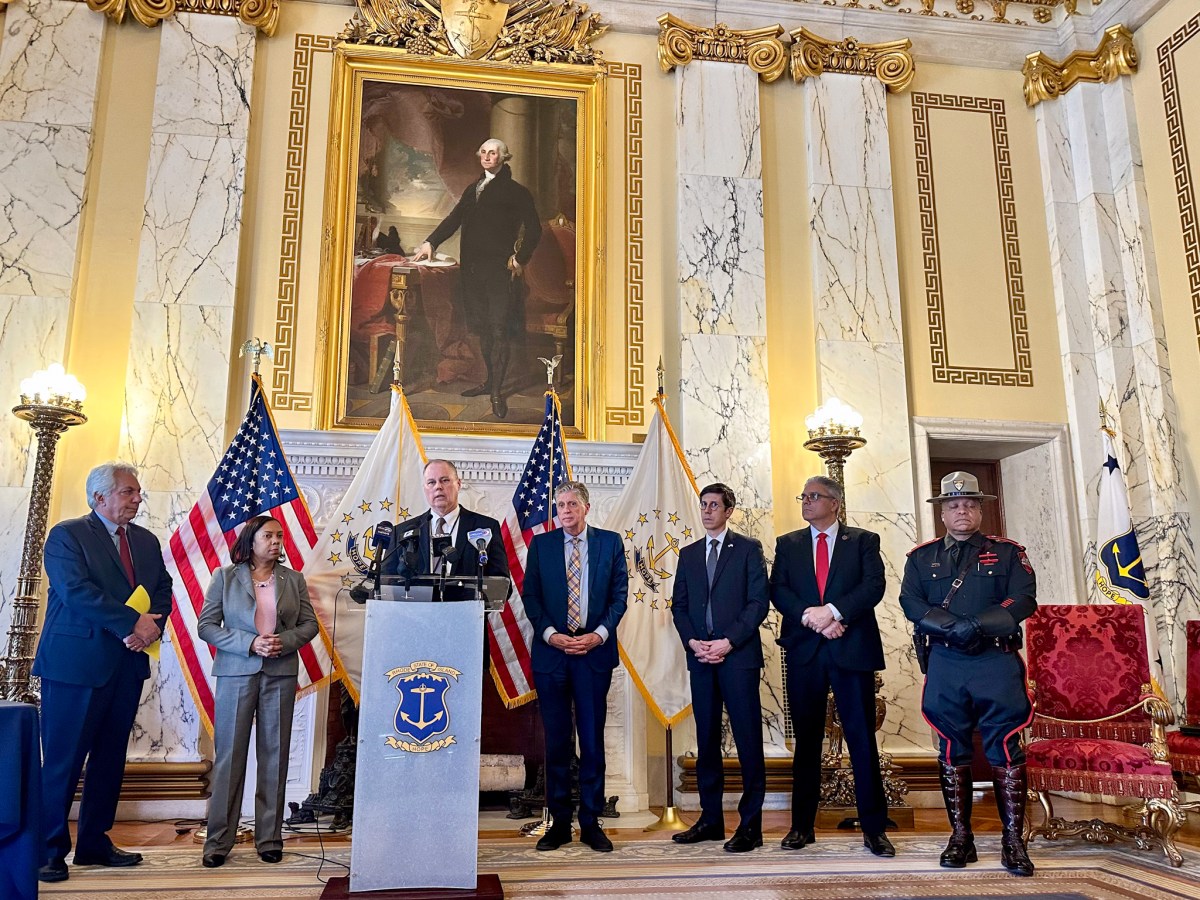 Multiple male government officials and one female government official stand behind a podium and in front of an ornate wall with a large oil painting. The room they are in has very high ceilings.