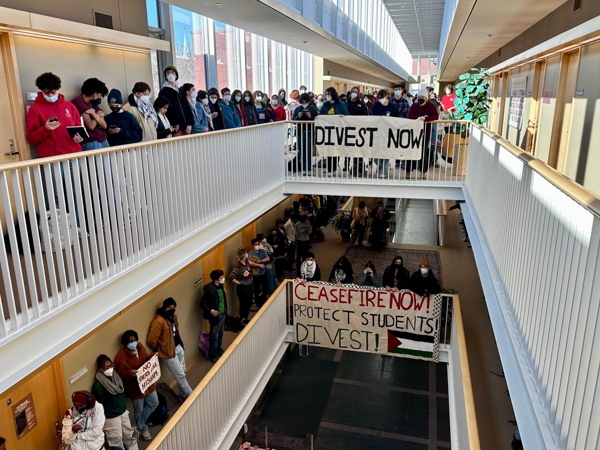 Masked Brown University students in a hallway waiting with large banners that say Divest Now and Ceasefire Now, Protect Students Divest with a Palestinian flag